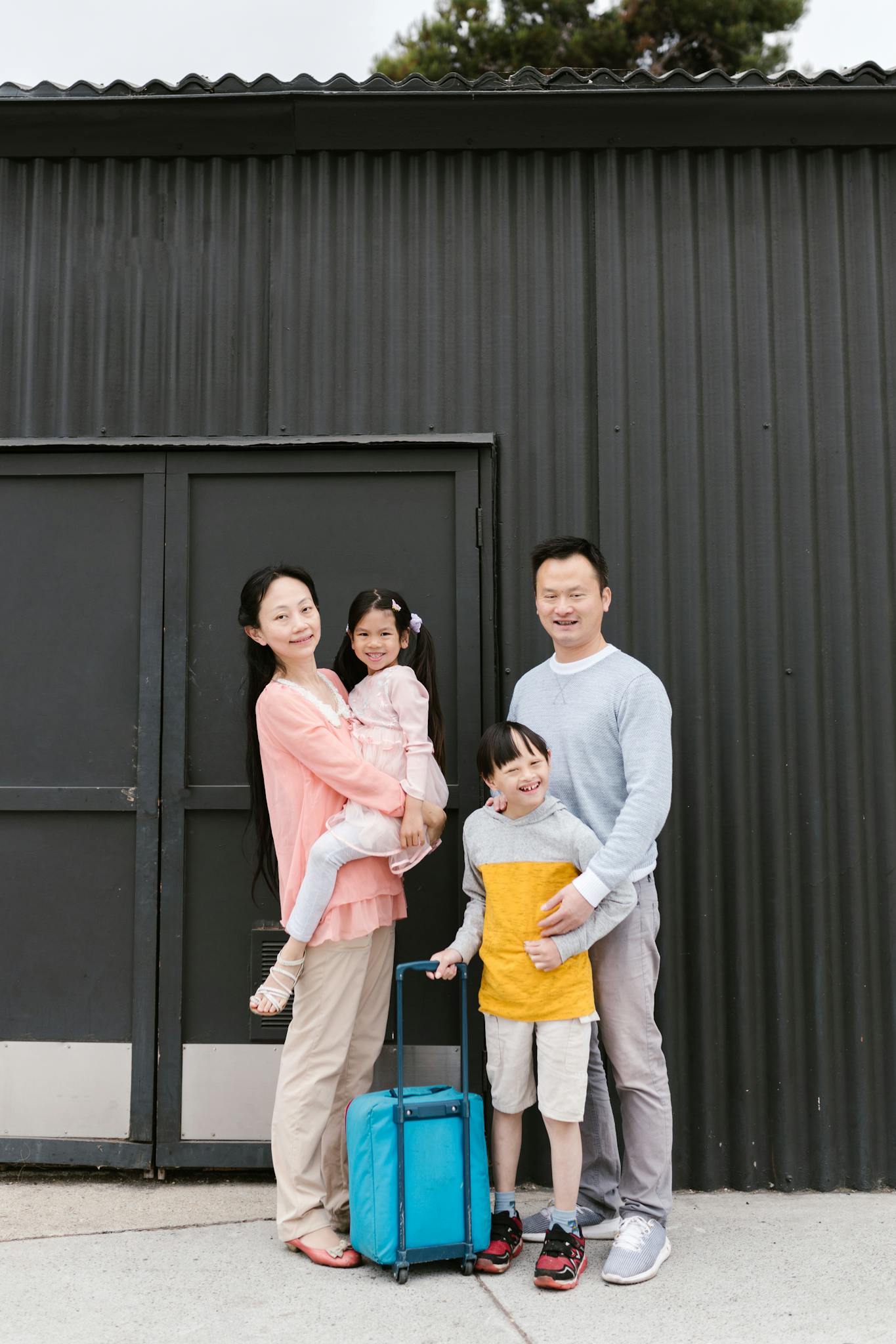 Asian family with two kids, ready for travel, smiling outdoors with luggage.